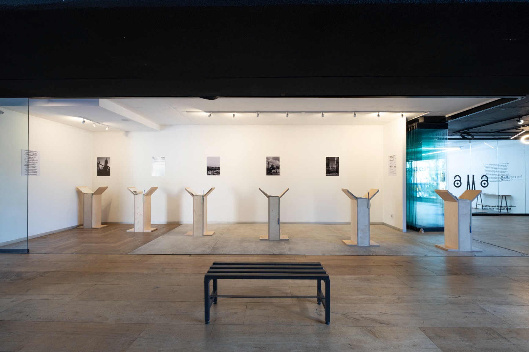 Photo of artwork. Artwork is a series of 6 timber podiums seen from afar in a gallery with photograohs on the wall.