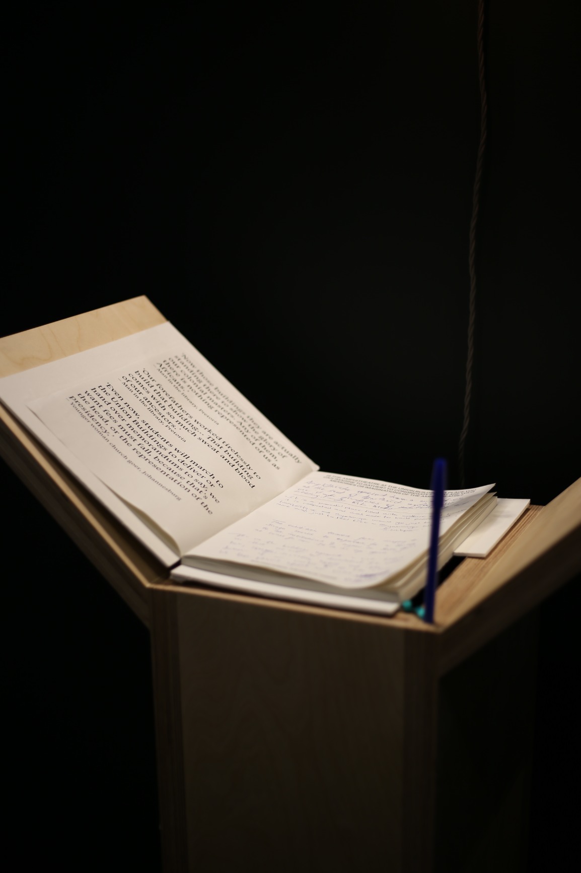 Photo of artwork fragment. Artwork is a series of 6 timber podiums. One podium is seen in this photo with the book on top of podium. The background is darkly lit.