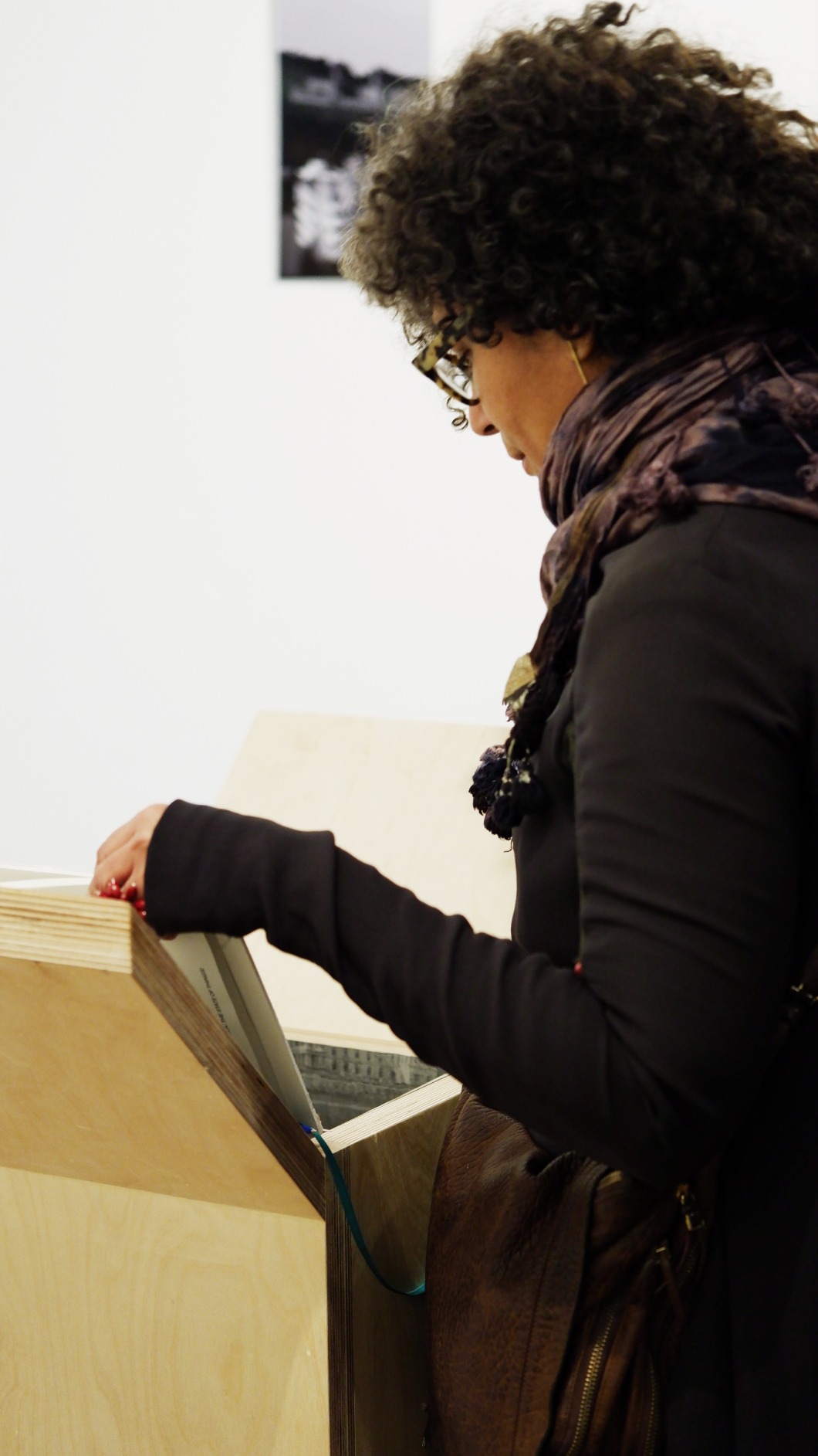 Photo of artwork fragment being engaged with by visitor. Artwork is a series of 6 timber podiums. One podium seen in this photo with woman peeking into podium interior, flipping the page of the book to open the interior.