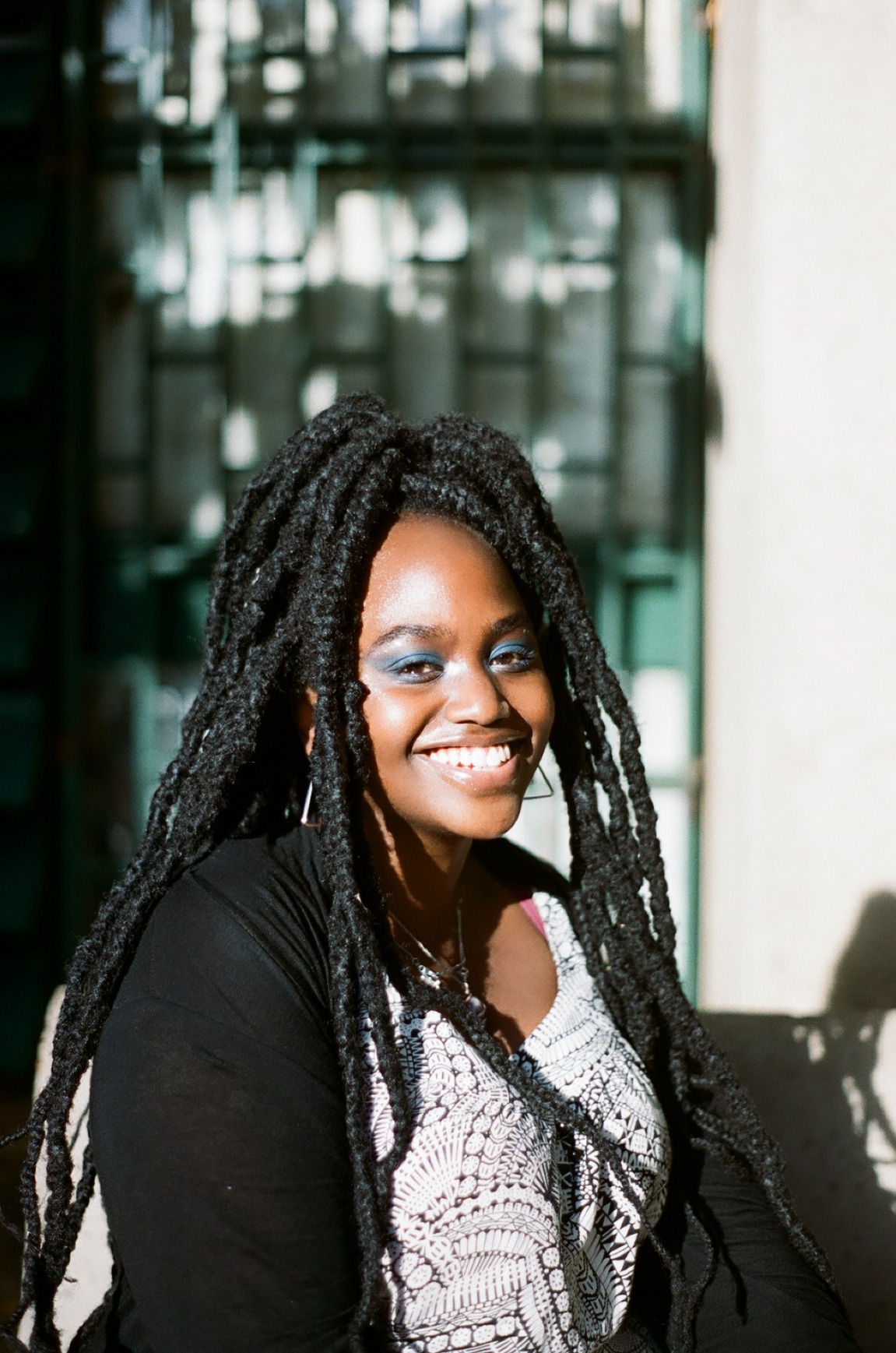 Photo portrait of young African woman smiling with braids and bright blue eye make-up, a white top and black blouse over it.