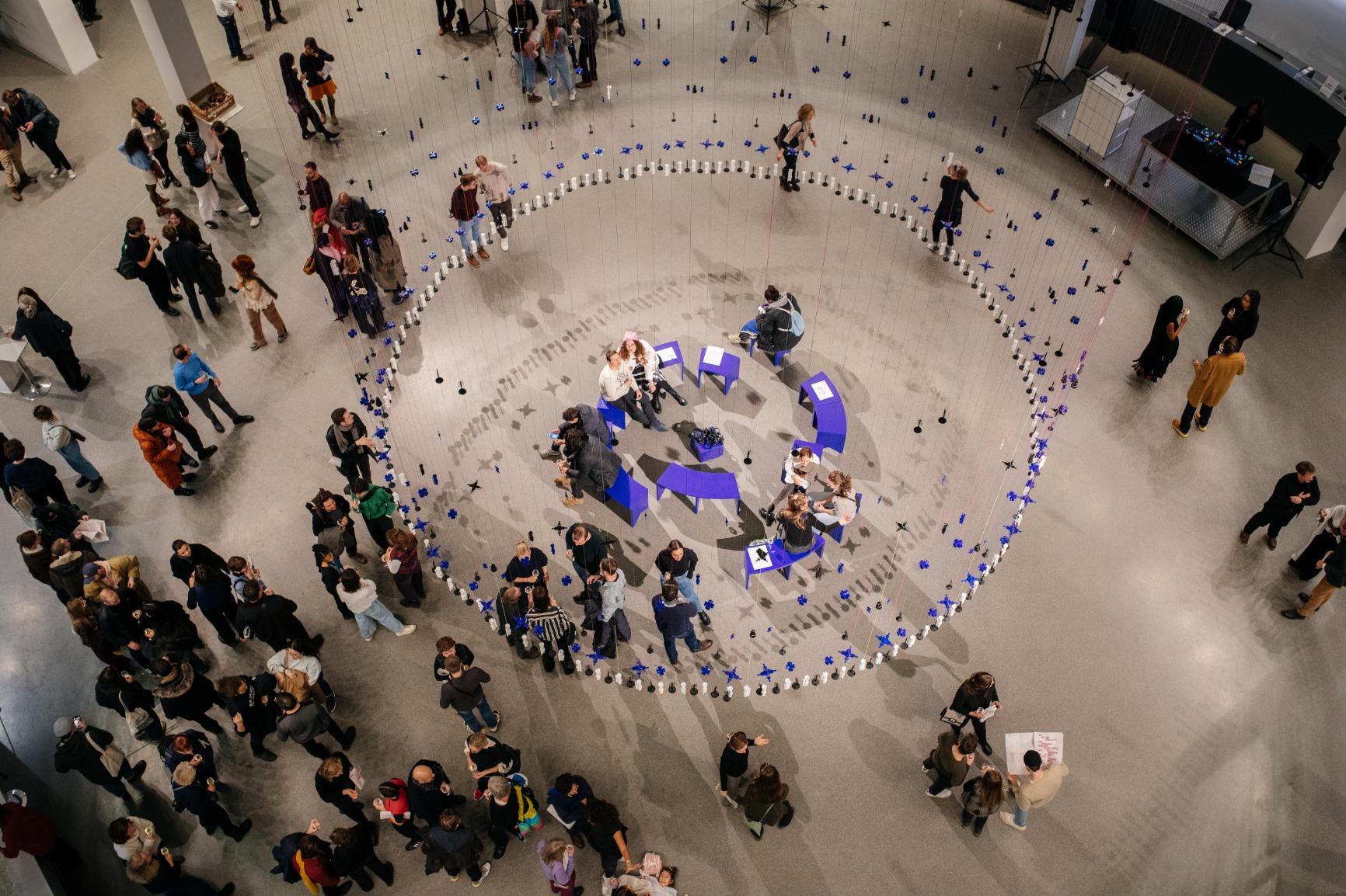 Photo of artwork. Artwork is a transparent beaded hanging curtain of 16m in a circular form creating an interior. The interior has blue benches that when put together form a circle. The curtain beads are blue, white and black. The strings are blue and pink. There are people sitting on the bench listening with earphones to a soundscape. There are people standing outside and inside the curtain. The image is taken during the opening evening.