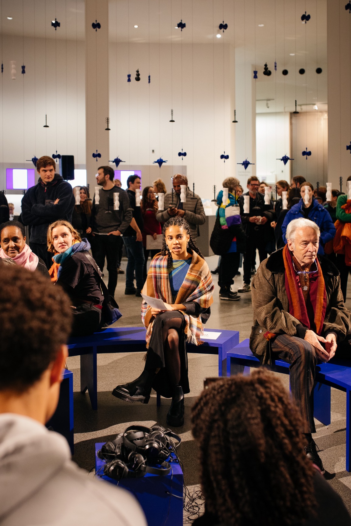Photo of artwork. Artwork is a transparent beaded hanging curtain of 16m in a circular form creating an interior. The interior has blue benches that when put together form a circle. The curtain beads are blue, white and black. The strings are blue and pink. There are people sitting on the bench listening with earphones to a soundscape. There are people standing outside and inside the curtain. The image is taken during the opening evening where Abdé is loudreading.