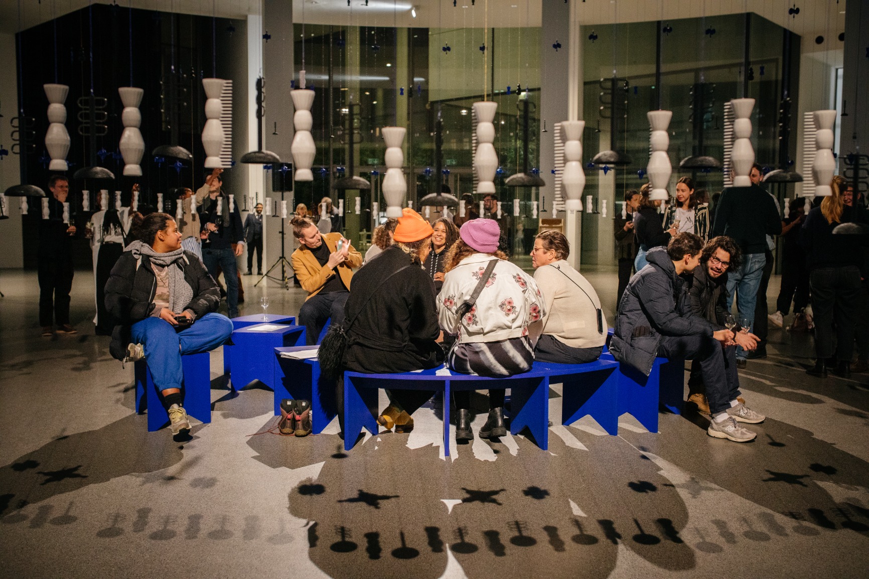 Photo of artwork. Artwork is a transparent beaded hanging curtain of 16m in a circular form creating an interior. The interior has blue benches that when put together form a circle. The curtain beads are blue, white and black. The strings are blue and pink. There are people sitting on the bench listening with earphones to a soundscape. There are people standing outside and inside the curtain. The image is taken during the opening evening.
