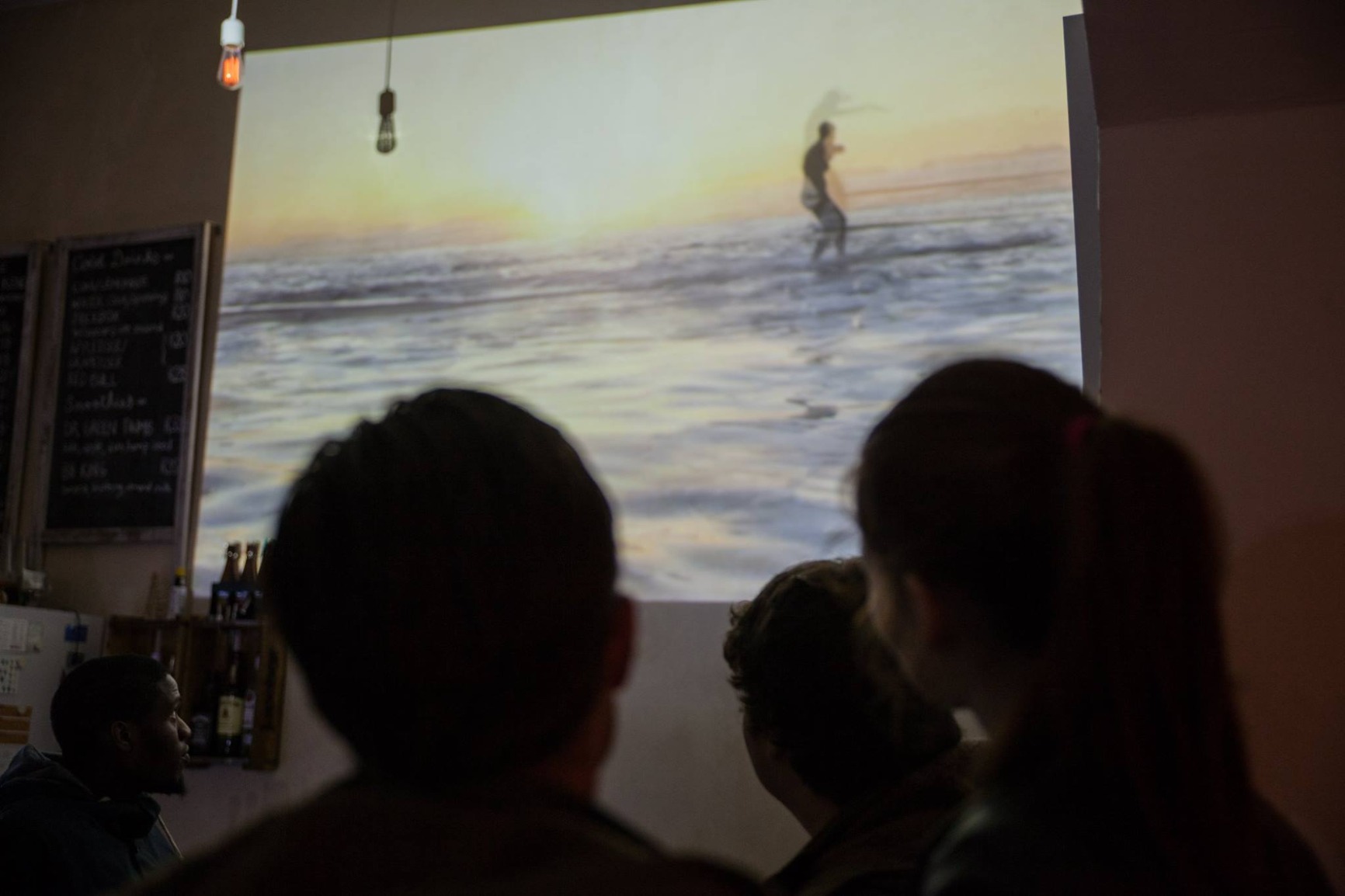 Photo of Ode to An Architect film screening showing the silhouettes of three people in the audience watching the film on a projector. The scene still shows a figure dancing at the ocean.