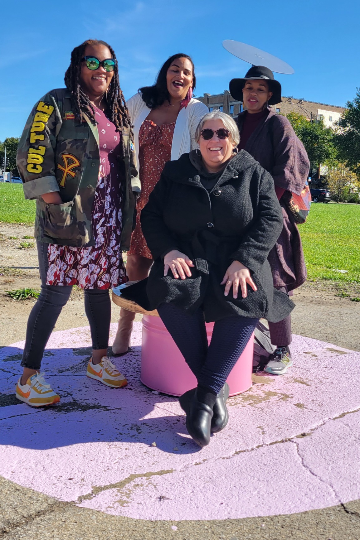 Photo showing pavilion pink bench with four invited poets, one sitting and the other three standing. All women are people of colour and smiling.