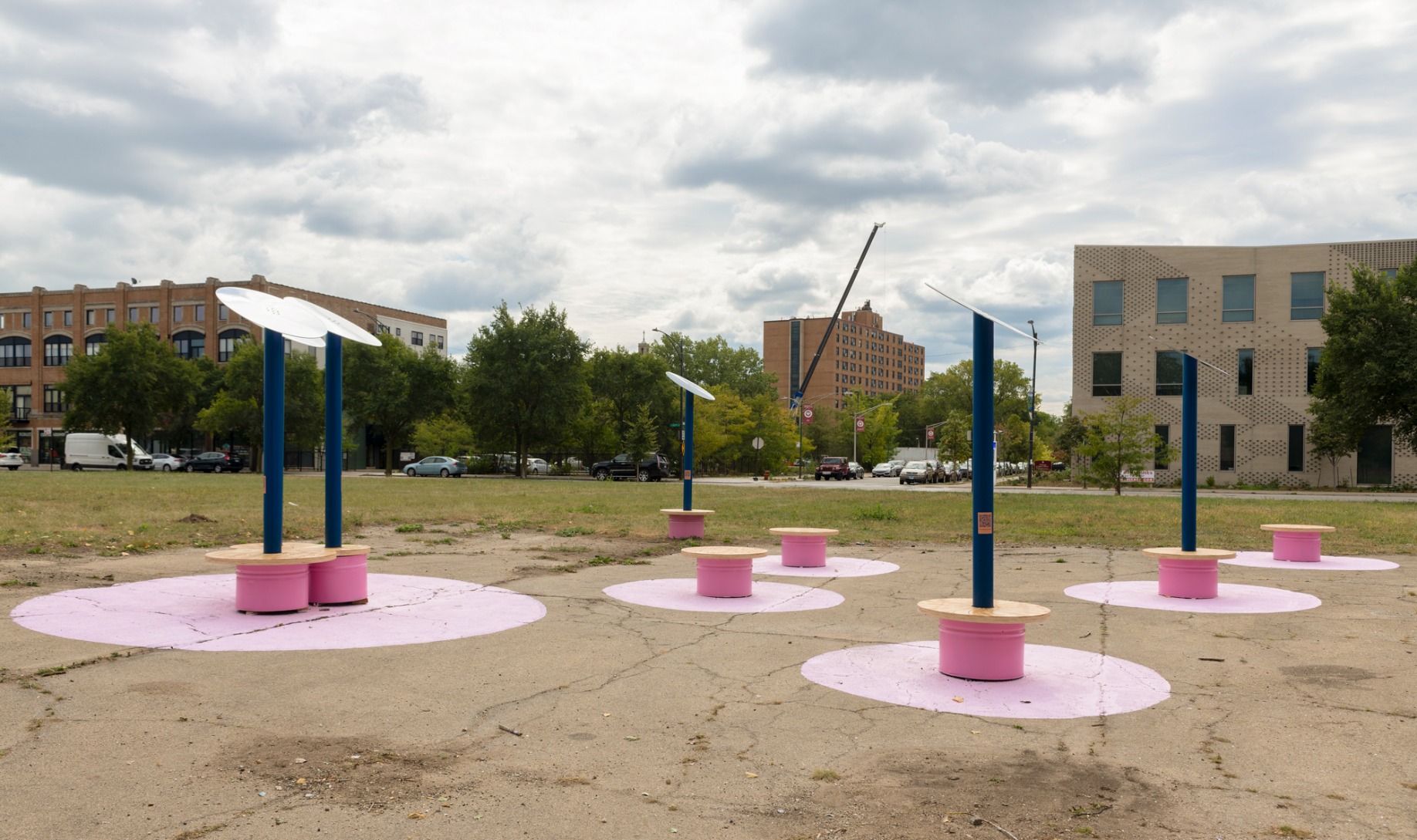 Photo showing pavilion from afar. Reflective disks are held on top of blue poles which are fixed into pink benches with timber tops. The full lawn is visible and neighbouring buildings. The ground is concrete with pink circles painted onto it where a bench appears.