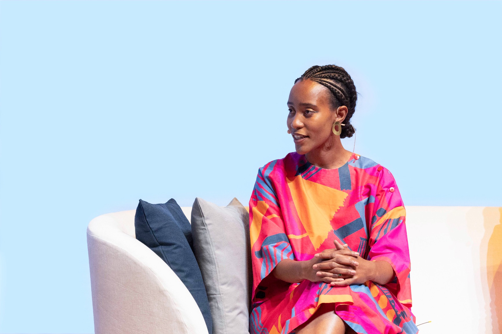 Photo of Khensani Jurczok-de Klerk sitting on a white couch speaking, wearing a bright colourful dress. The colours are pink, orange and blue. She has braids.
