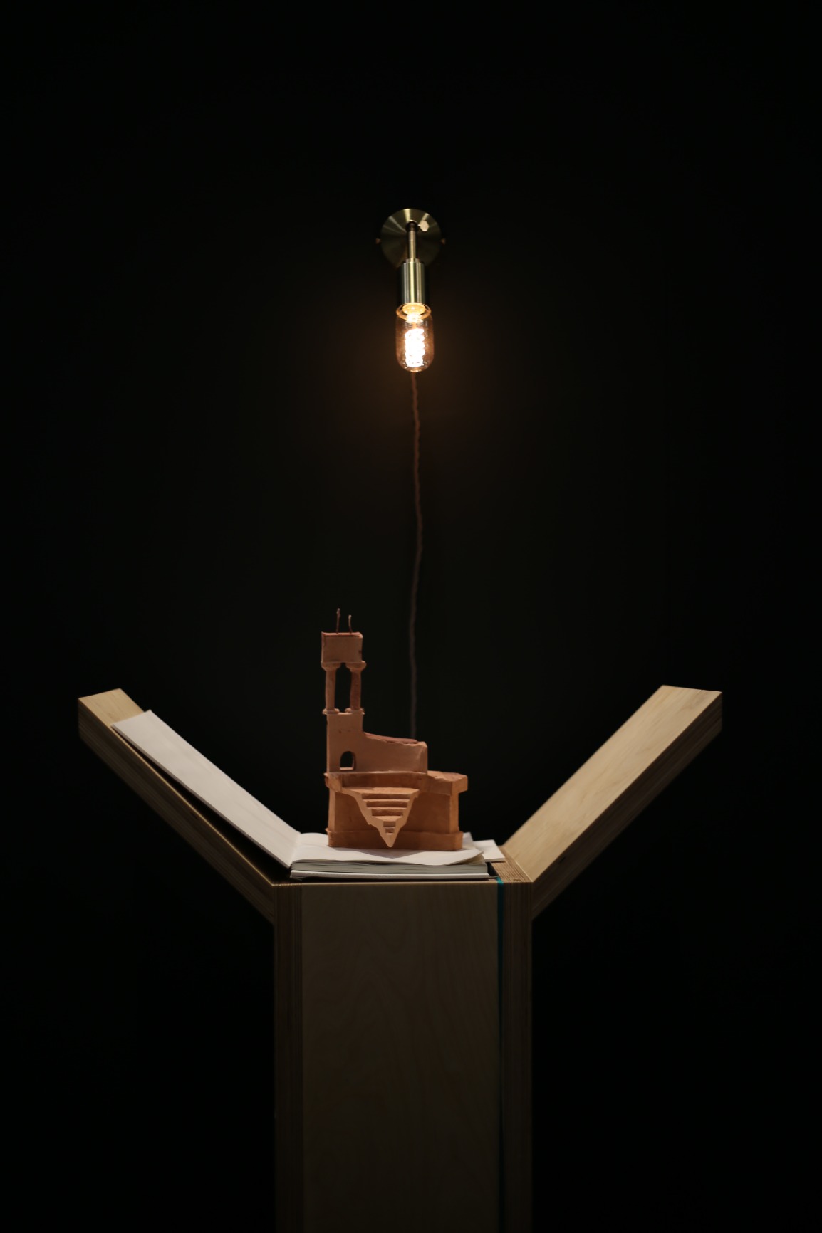Photo of artwork fragment. Artwork is a series of 6 timber podiums. One podium is seen in this photo with clay model of ampitheatre sitting on top of book on top of podium. The background is darkly lit.