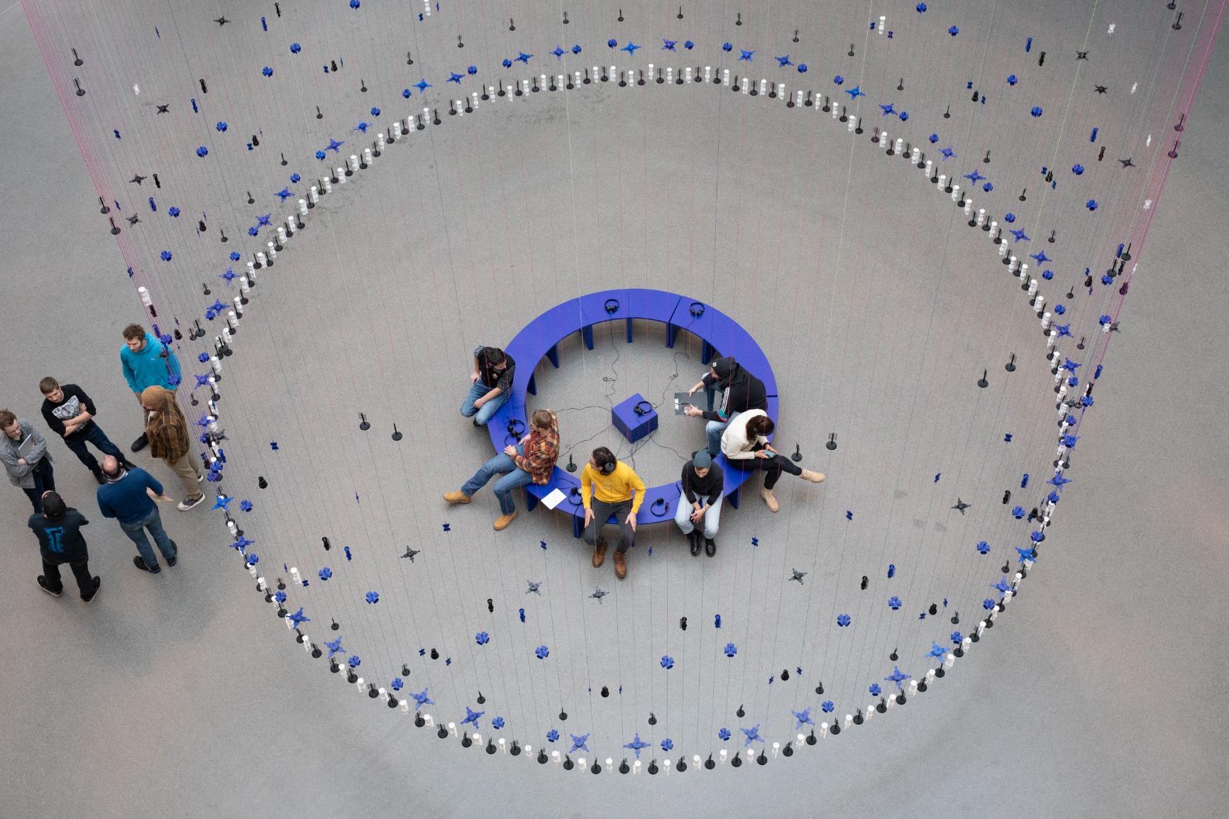 Photo of artwork. Artwork is a transparent beaded hanging curtain of 16m in a circular form creating an interior. The interior has blue benches that when put together form a circle. The curtain beads are blue, white and black. The strings are blue and pink. There are people sitting on the bench listening with earphones to a soundscape. There are people standing outside of the curtain. The image is taken from above. 