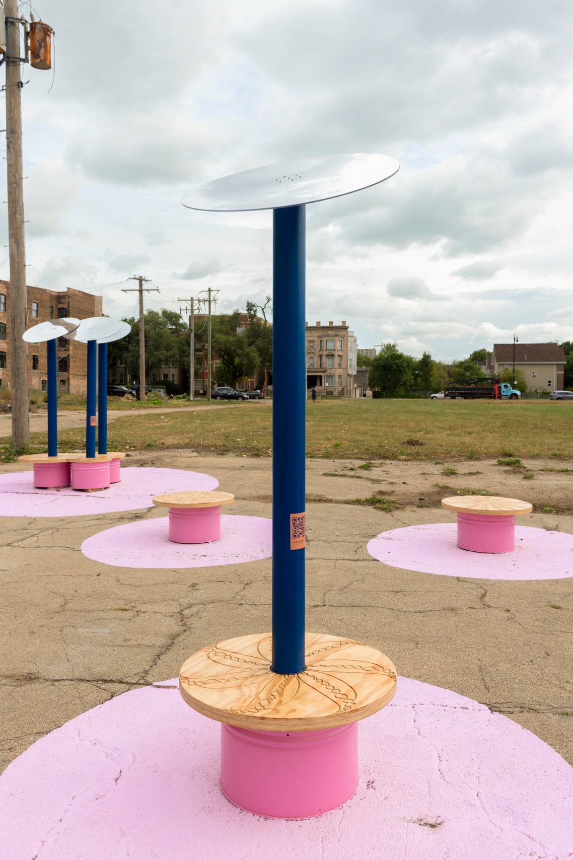 Photo showing pavilion. Reflective disks are held on top of blue poles which are fixed into pink benches with timber tops. There are pink rings painted on the ground where a bench appears. There are QR codes on each pole.