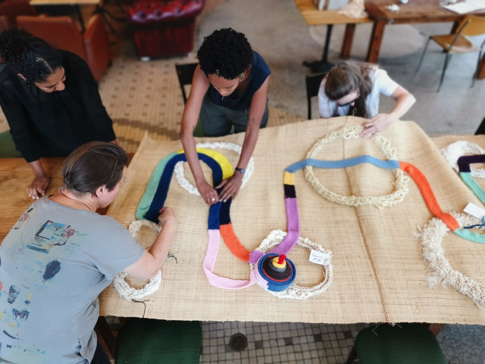 Photo of Matri-Archi team members and collaborators sewing items onto the tapestry. The tapestry is a rafia-base with colourful woolen swirls interrupted by rings of various textile materials.
